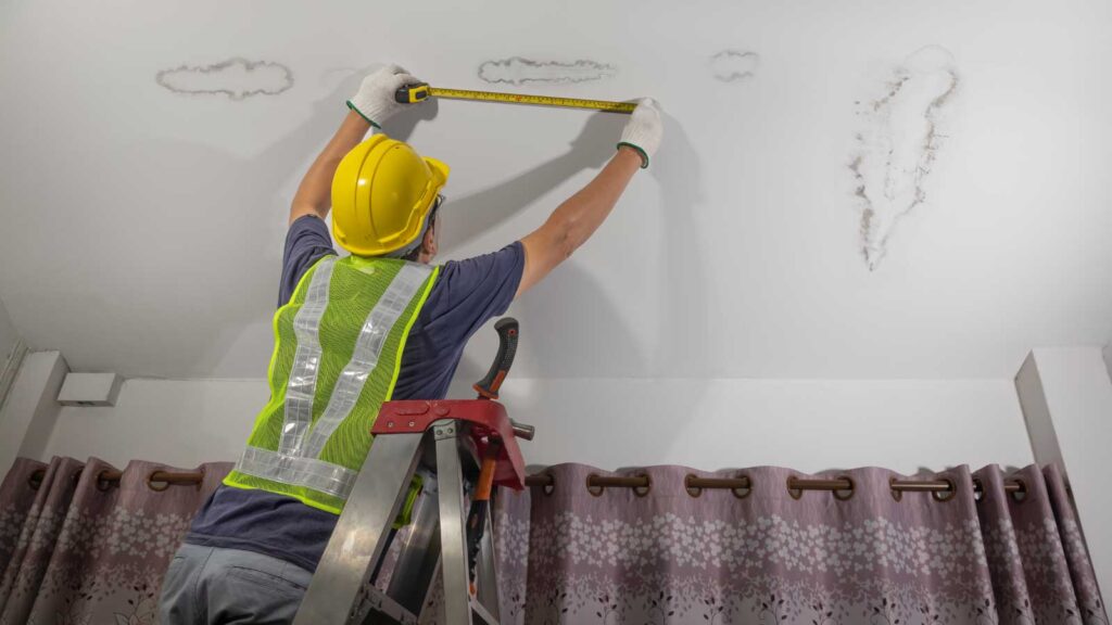 Worker measures ceiling water damage with a tape measure.