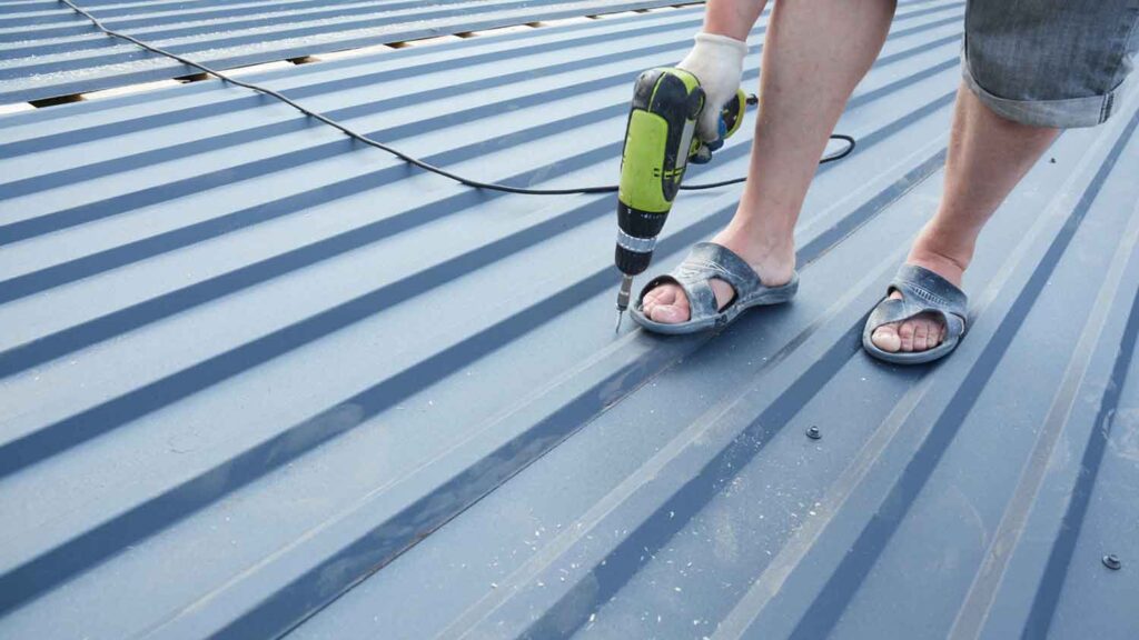 A worker drills screws into a metal roof panel.