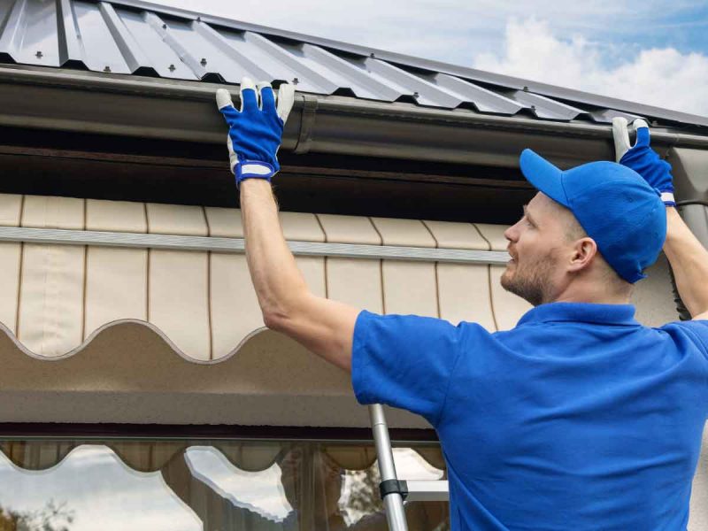 Man in blue uniform inspecting roof gutter from a ladder.