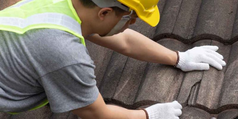 Roofer in safety gear repairing broken roof tiles with a hammer.