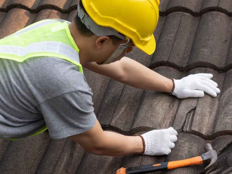 Roofer in safety gear repairing broken roof tiles with a hammer.